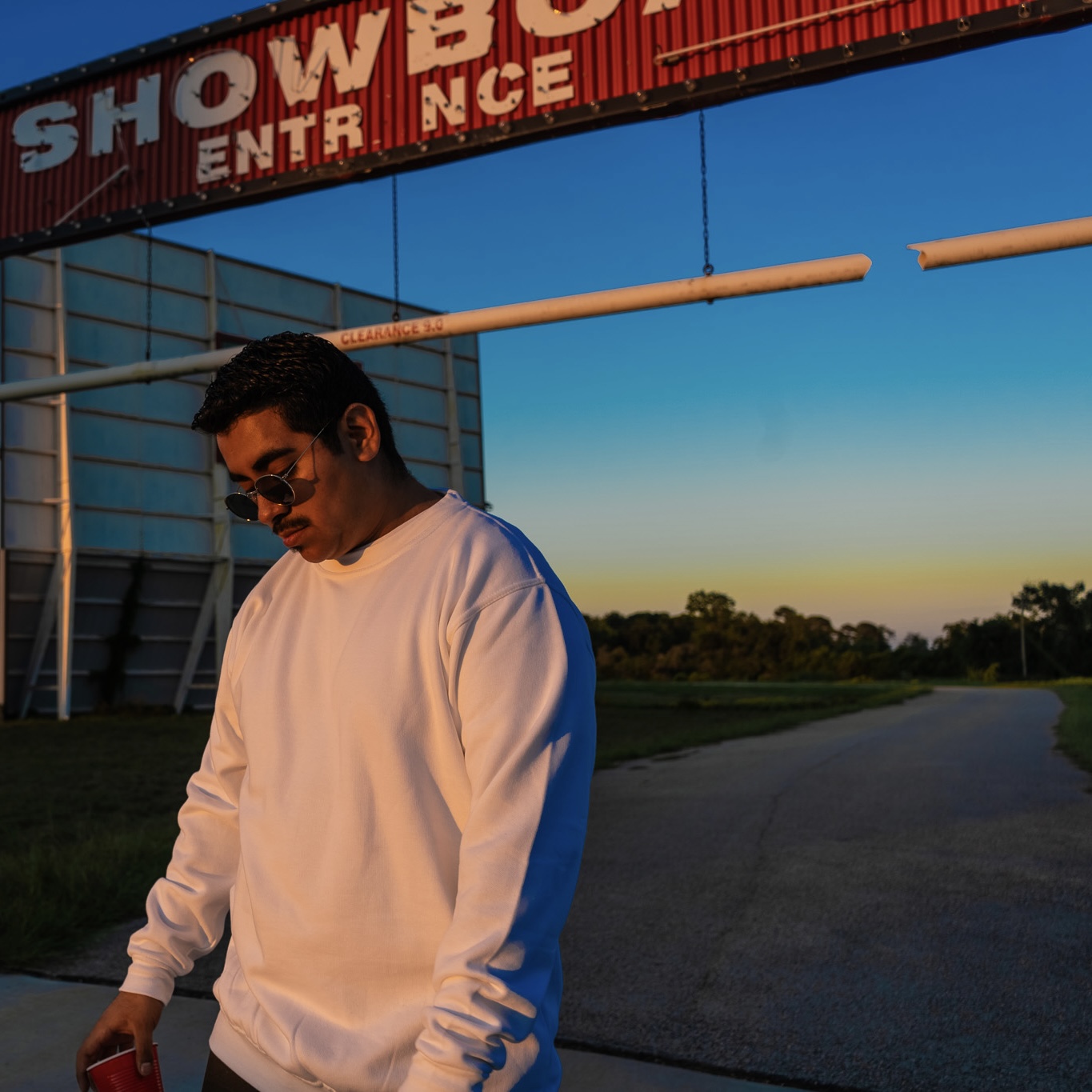 Young man with sunglasses wearing a white sweatshirt standing outdoors near a building and road, sunset sky in background.