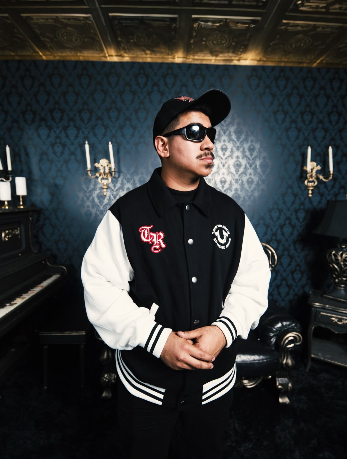 Young man wearing sunglasses, cap, and varsity jacket standing in a room with dark blue wallpaper and chandeliers.