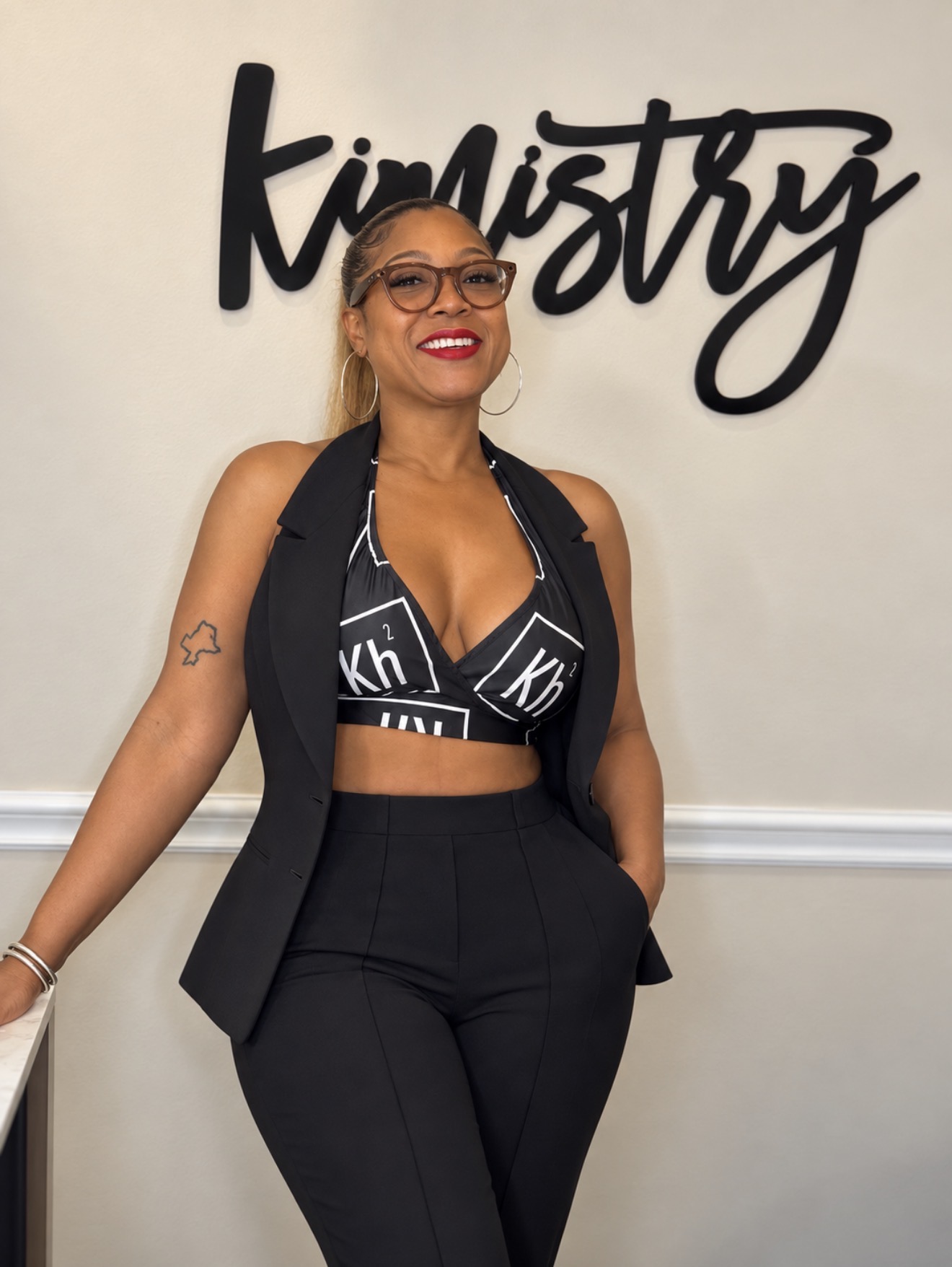 Woman with glasses and blonde hair smiling, standing in front of a wall with black script text, wearing a black outfit.