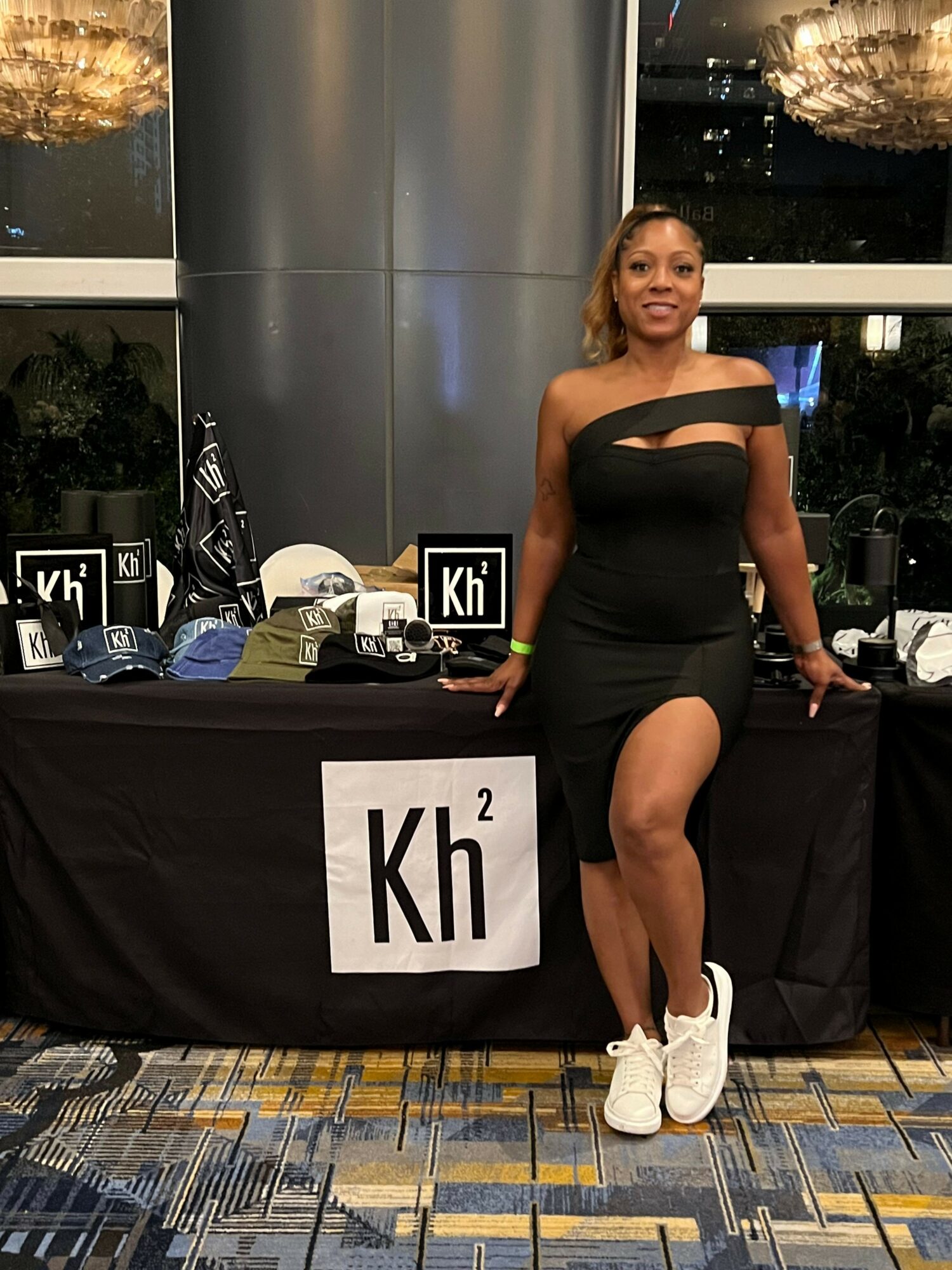 Woman in black dress and white sneakers standing next to a table with promotional materials, in an indoor setting.