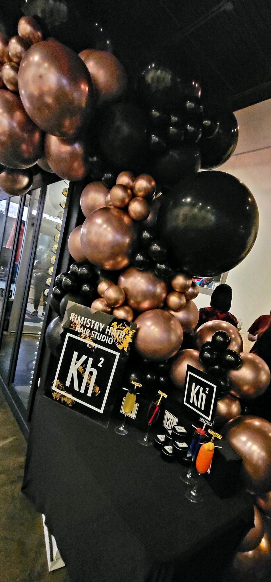 Balloon display with black, bronze, and gold balloons arranged vertically, with a table holding small decorations and signs, indoors.