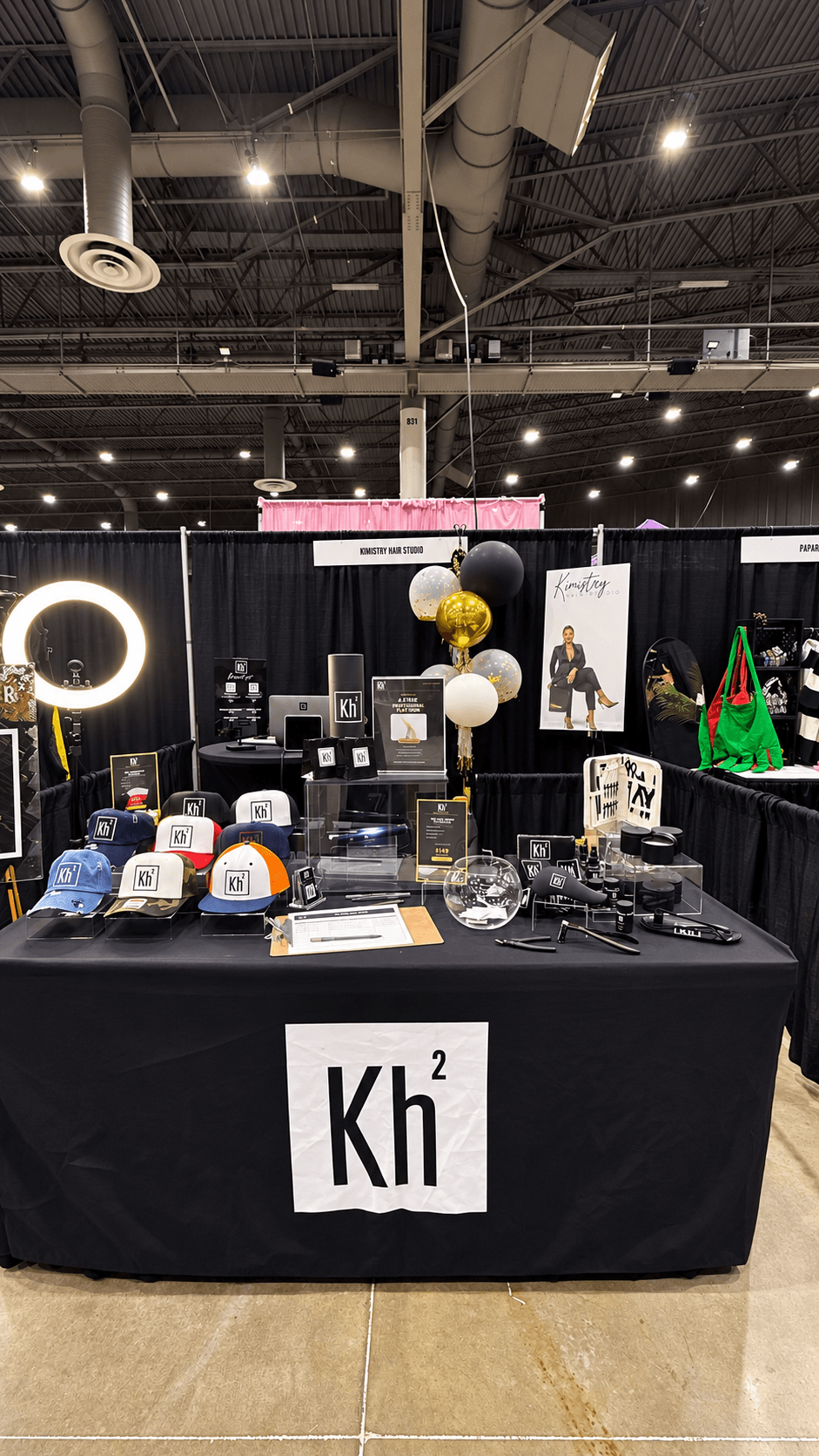 Display table with electronic items and decorations at an indoor event, with a black tablecloth and a sign reading 'Kh' in front.