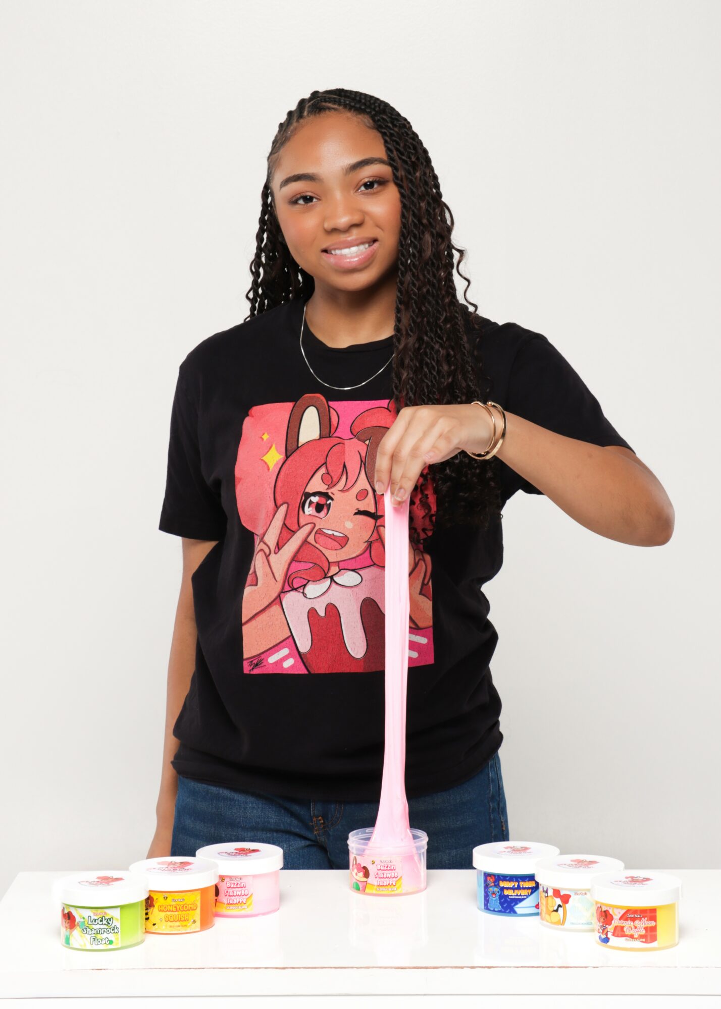 Young woman with long braided hair smiling, holding a pink slime tube, standing behind a table with colorful slime containers.