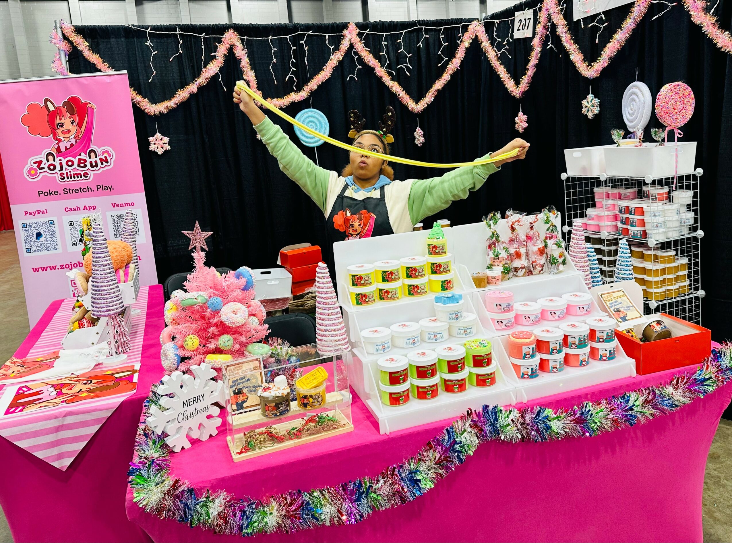 Person standing behind a colorful display table with sweets and decorations, pink tablecloth, and festive garlands, in a decorated party setting.