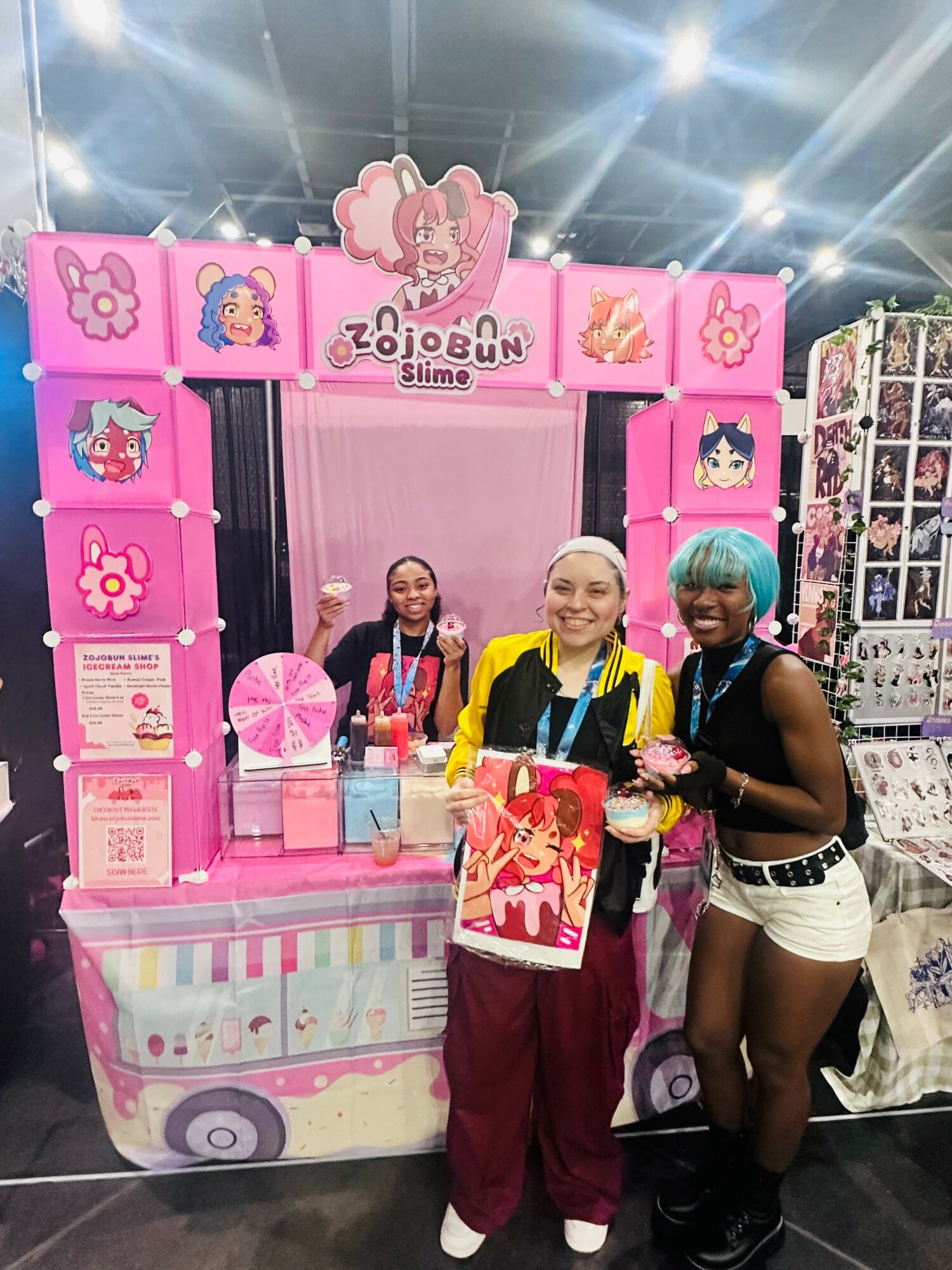 Three people stand in front of a pink Zojobun slime booth at a convention, smiling and holding slime containers.