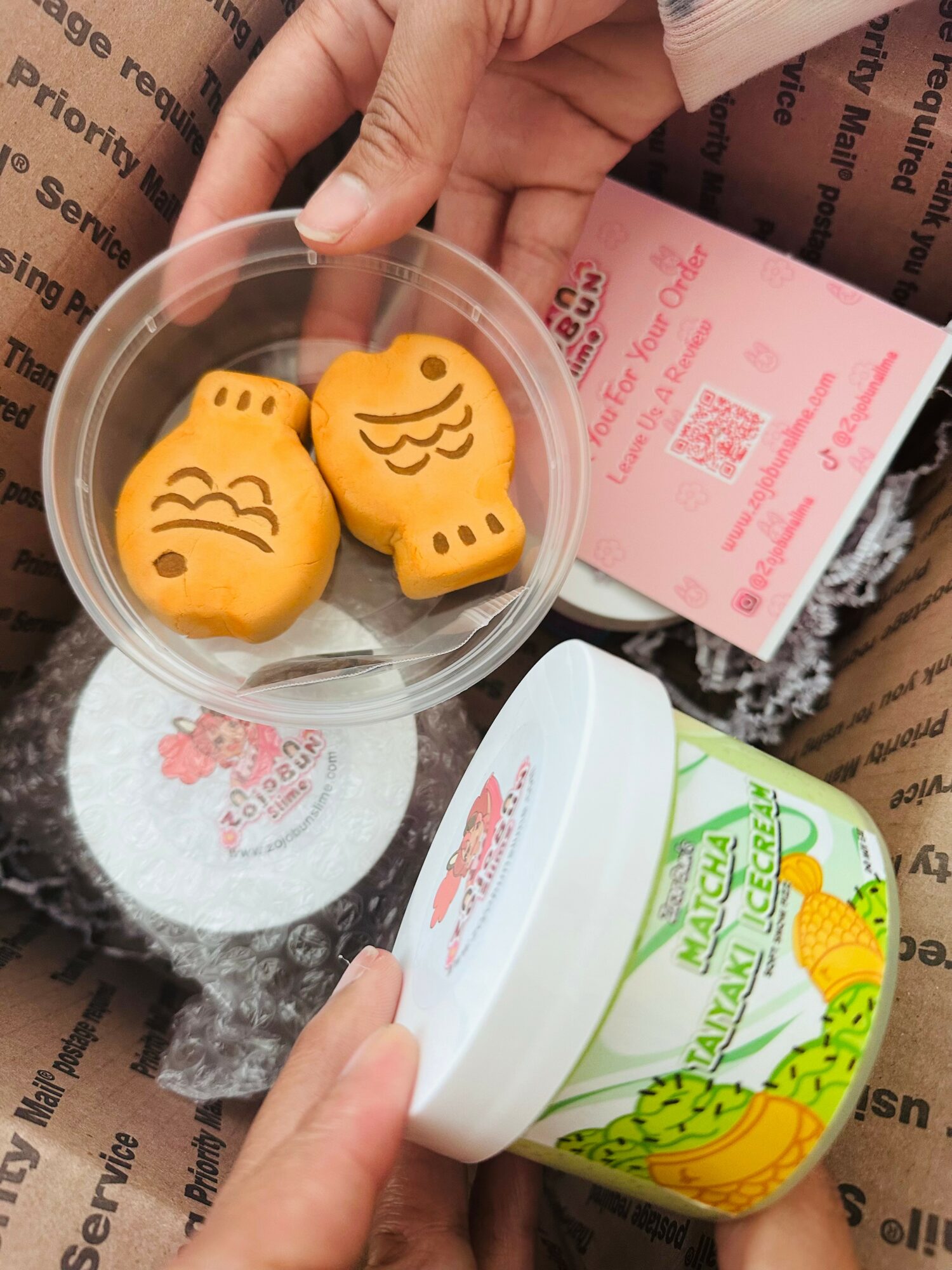 Person holding a clear container with three yellow animal-shaped cookies, and another container with a green and white label, on brown paper packaging.