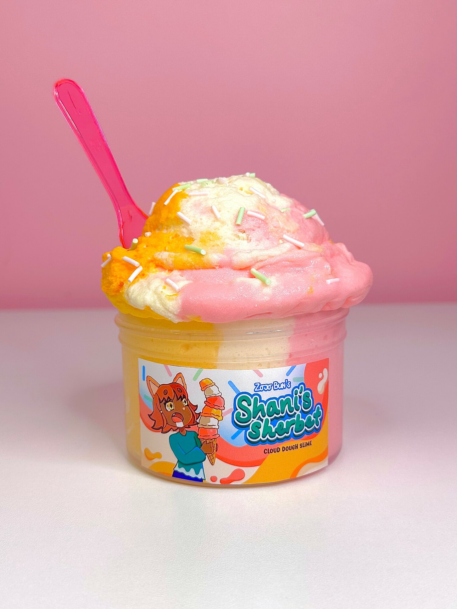 Colorful ice cream in a container with a pink spoon, pink background, and cartoon character on the label.