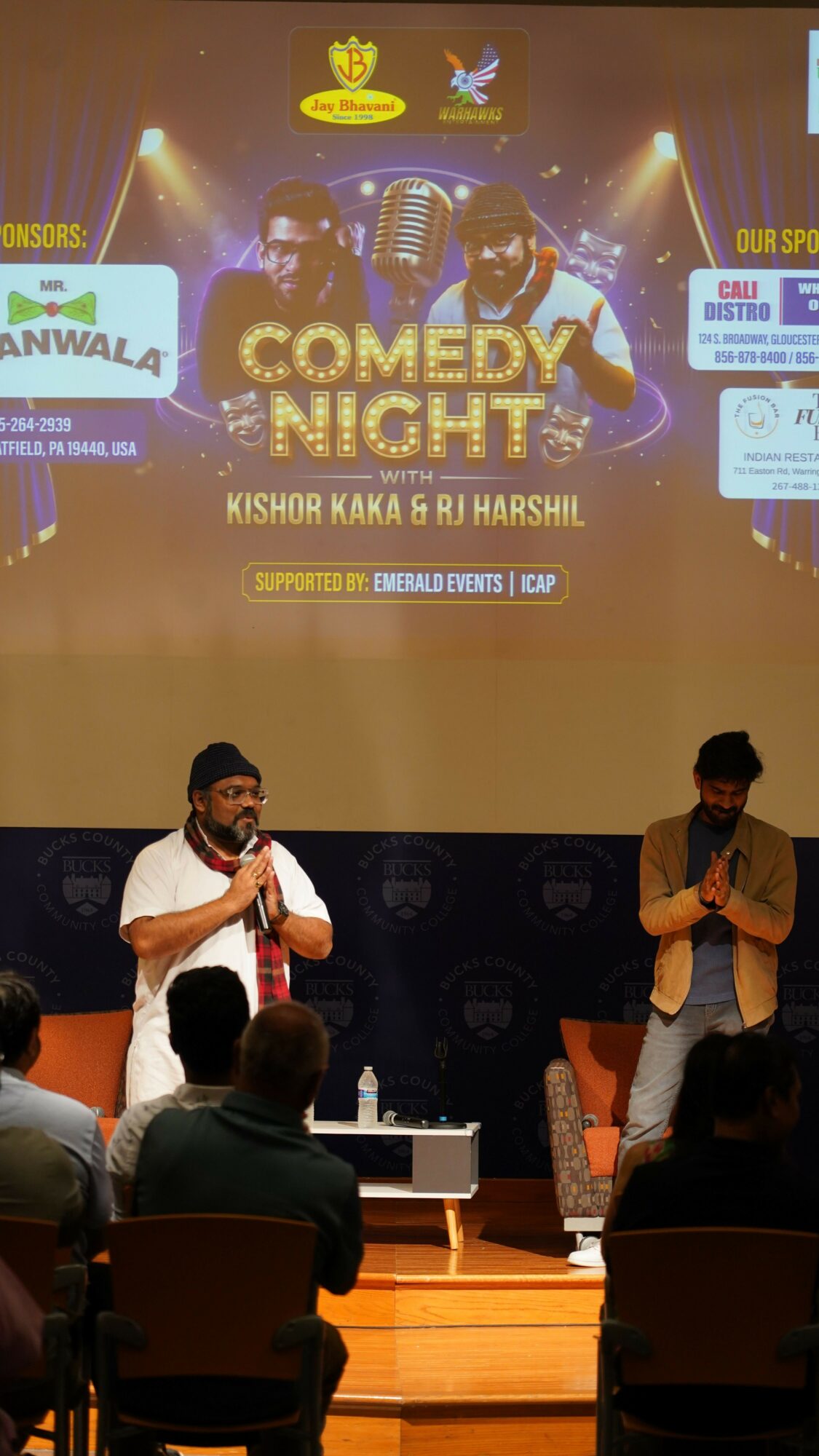 People on stage at comedy night event, with a large screen displaying event details behind them.