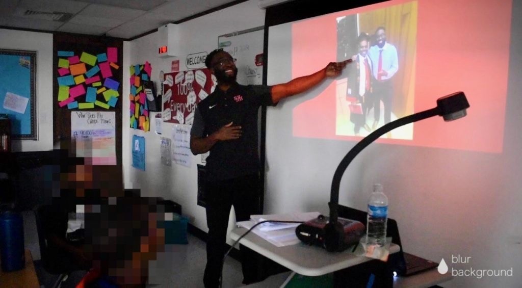 Person pointing at a projected presentation in a classroom with students, posters, and a desk with a water bottle.