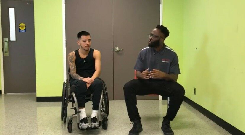 Two men sitting and talking in a room with green and gray walls, one in a wheelchair and the other on a bench.