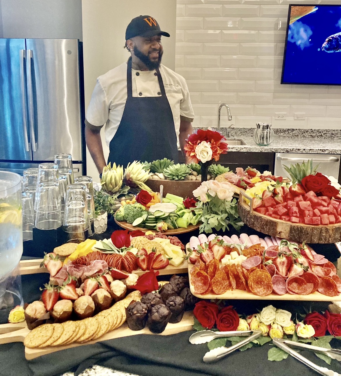 Man wearing a cap and apron stands behind a table with an assortment of fruit and flower arrangements, in a modern kitchen.