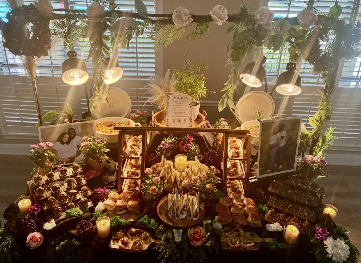 Decorative table with flowers, candles, and desserts, illuminated by overhead lights, with window blinds in the background.