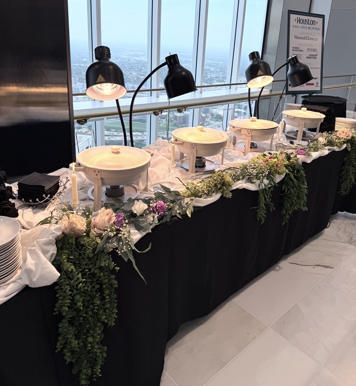 Buffet table with chafing dishes, floral arrangements, and lamps in a high-rise setting with city view, black tablecloth, and white marble floor.