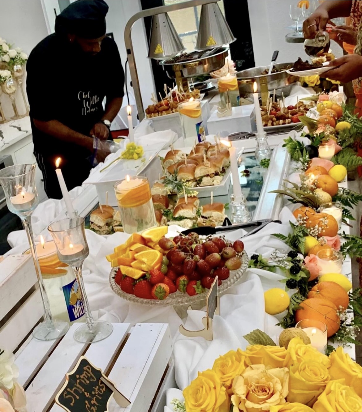 People serving food at a buffet table with fruits, desserts, and floral decorations, in a bright room.