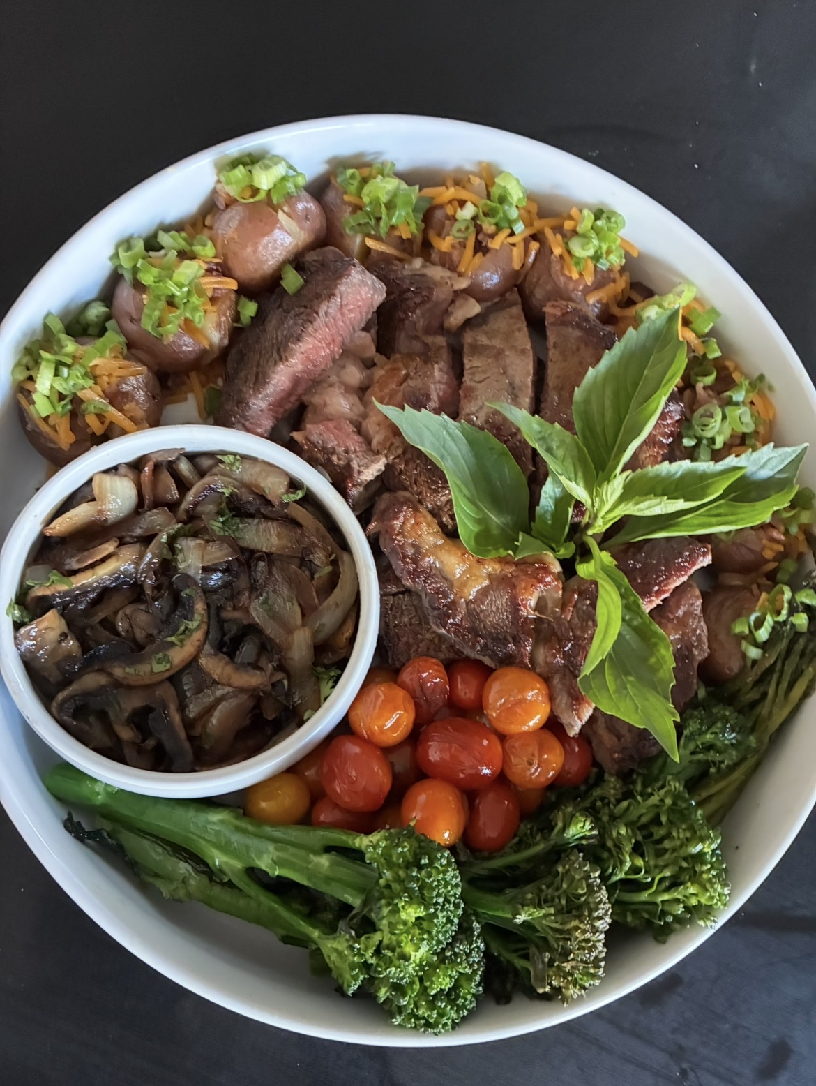 Assorted cooked vegetables, cherry tomatoes, and herbs on a white plate, with a small bowl of sautéed mushrooms and onions.