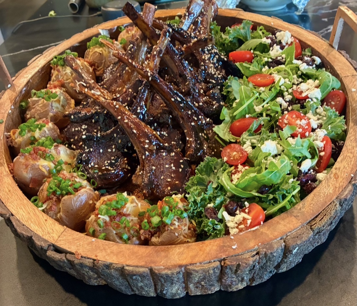 A wooden bowl with cooked lamb chops, a salad with cherry tomatoes, greens, and cheese, and roasted potatoes garnished with herbs.
