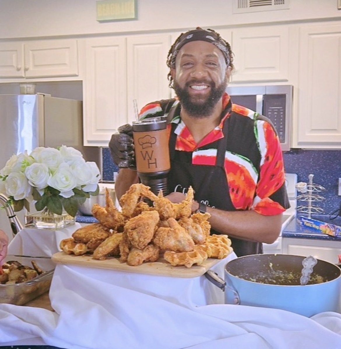 Man with a beard smiling, holding a drink, standing behind a table with fried chicken and waffles, in a kitchen.