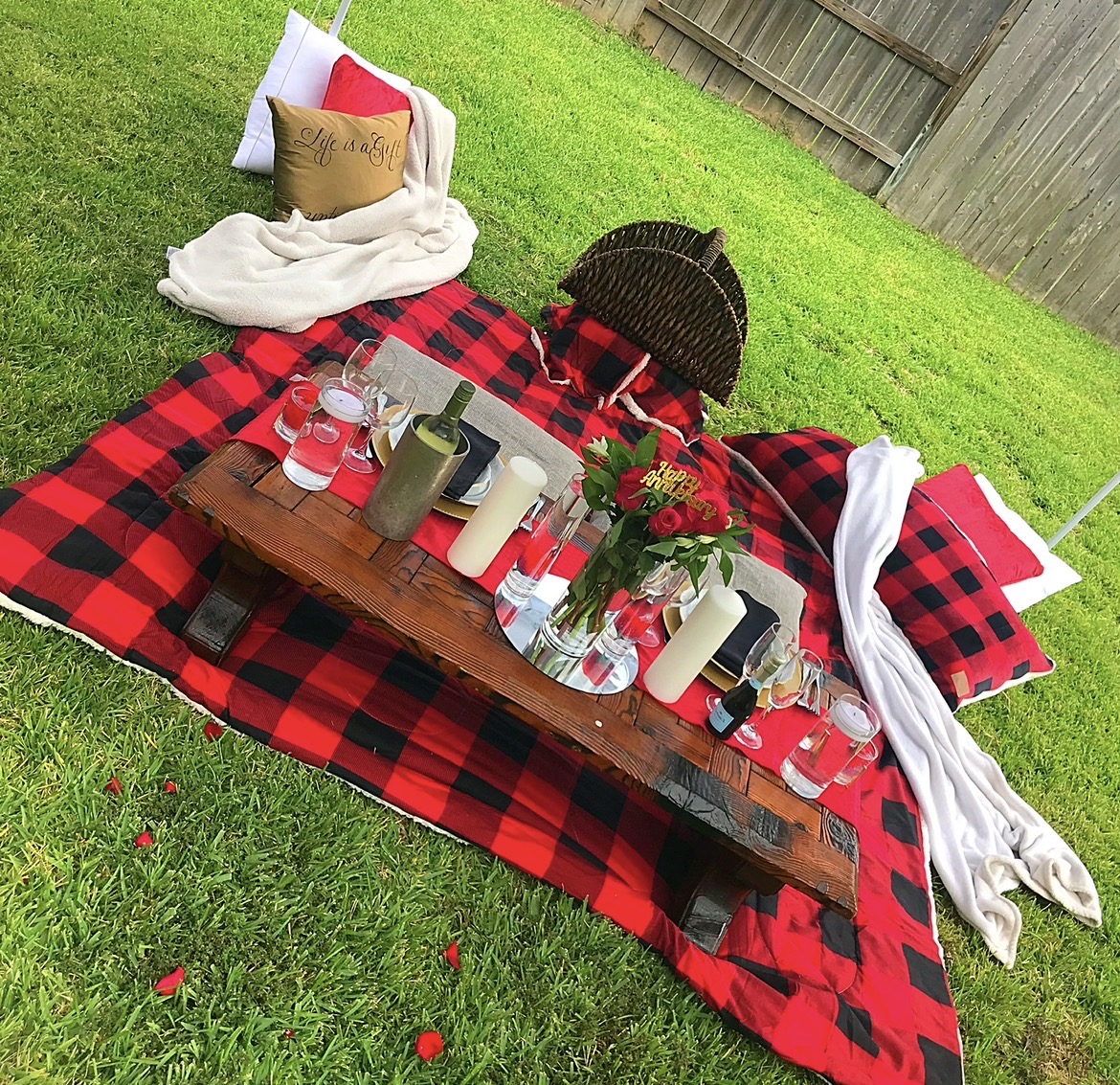 Picnic setup on a red and black checkered blanket with pillows, a wooden tray, candles, and flowers on grass.