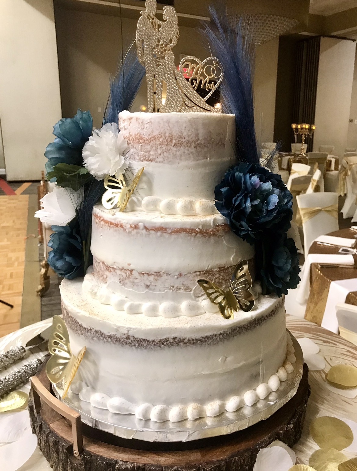 Three-tiered wedding cake decorated with blue and white flowers, gold butterflies, and a bride figurine topper, on a table in a banquet hall.