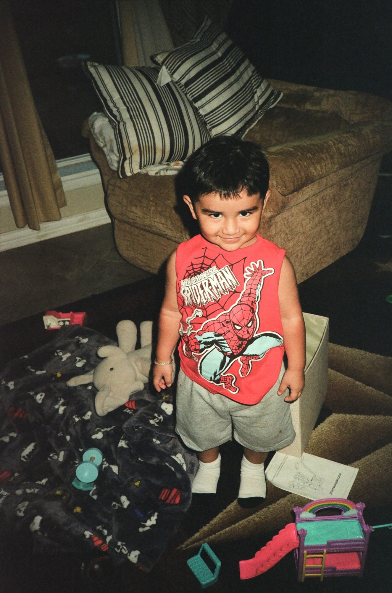 Young boy standing indoors near stairs, smiling, wearing a red shirt with a dinosaur graphic, shorts, and socks.