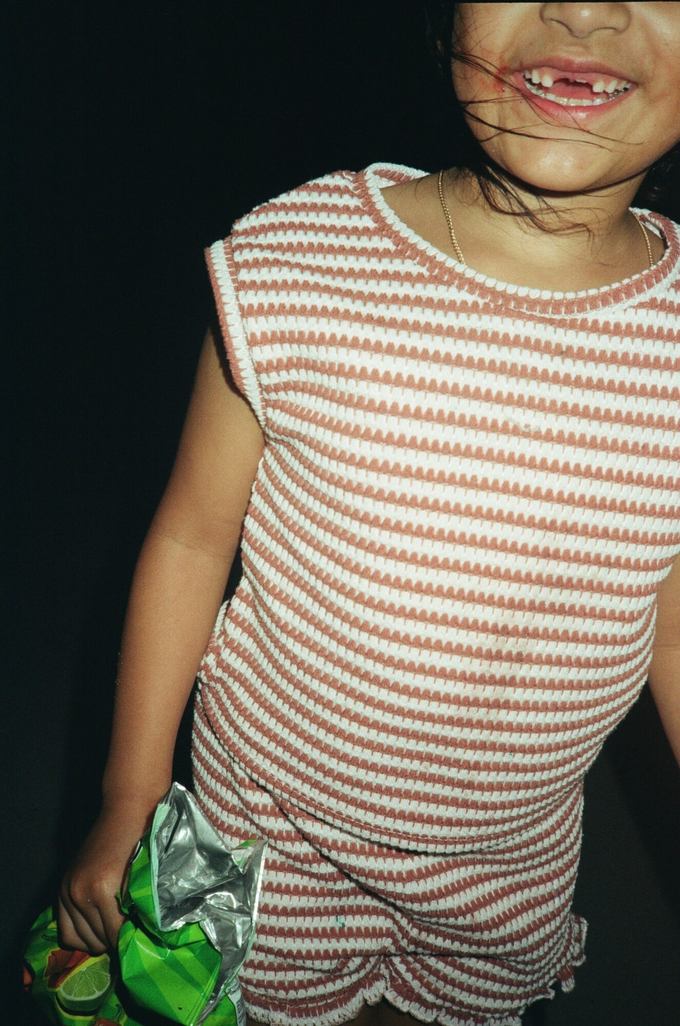 Child wearing a striped dress holding a shiny silver and green object, smiling with visible teeth, dark background.