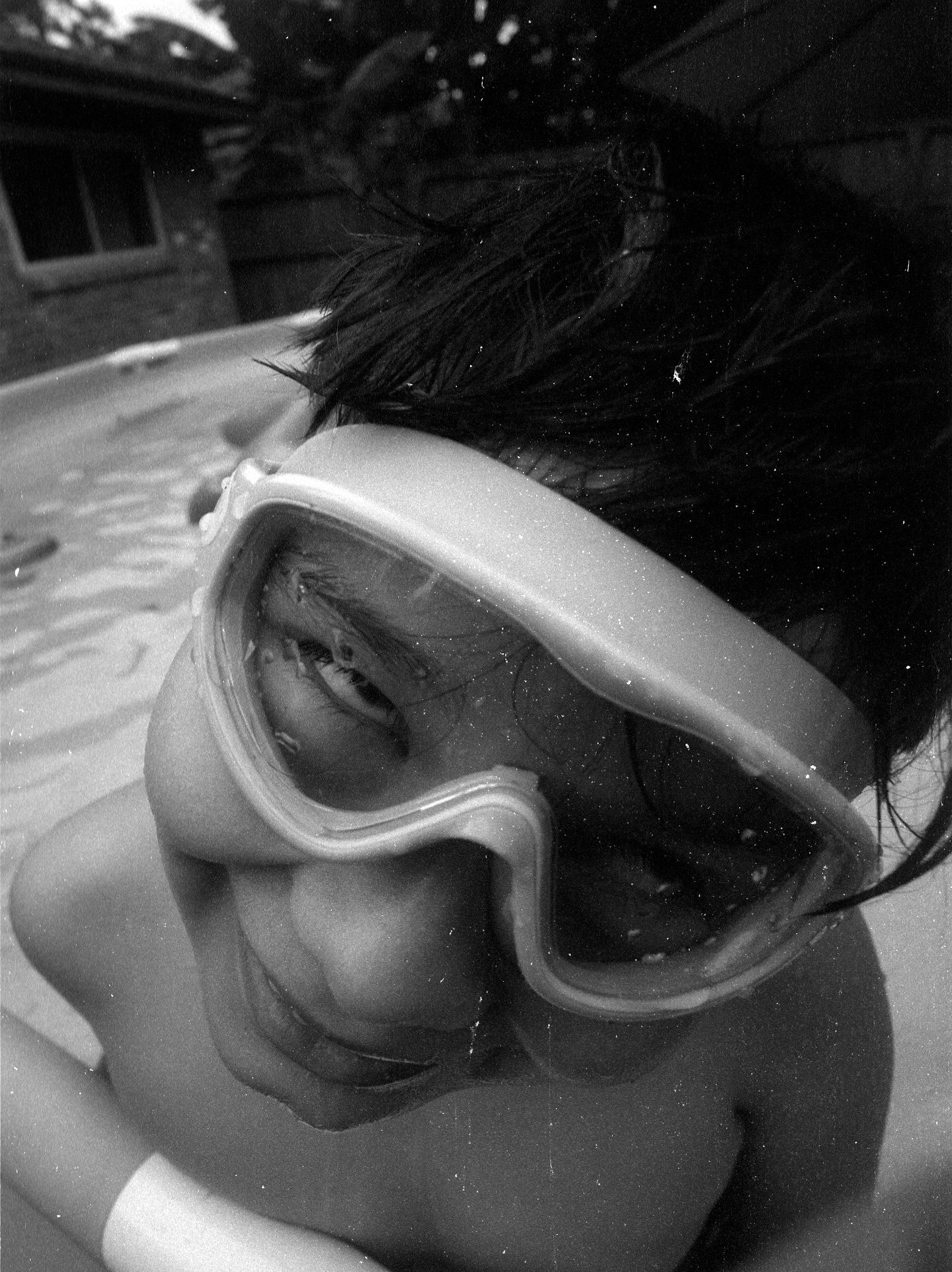 Child wearing goggles, smiling, outdoors, black and white photo, close-up, wet hair, background with house and trees.