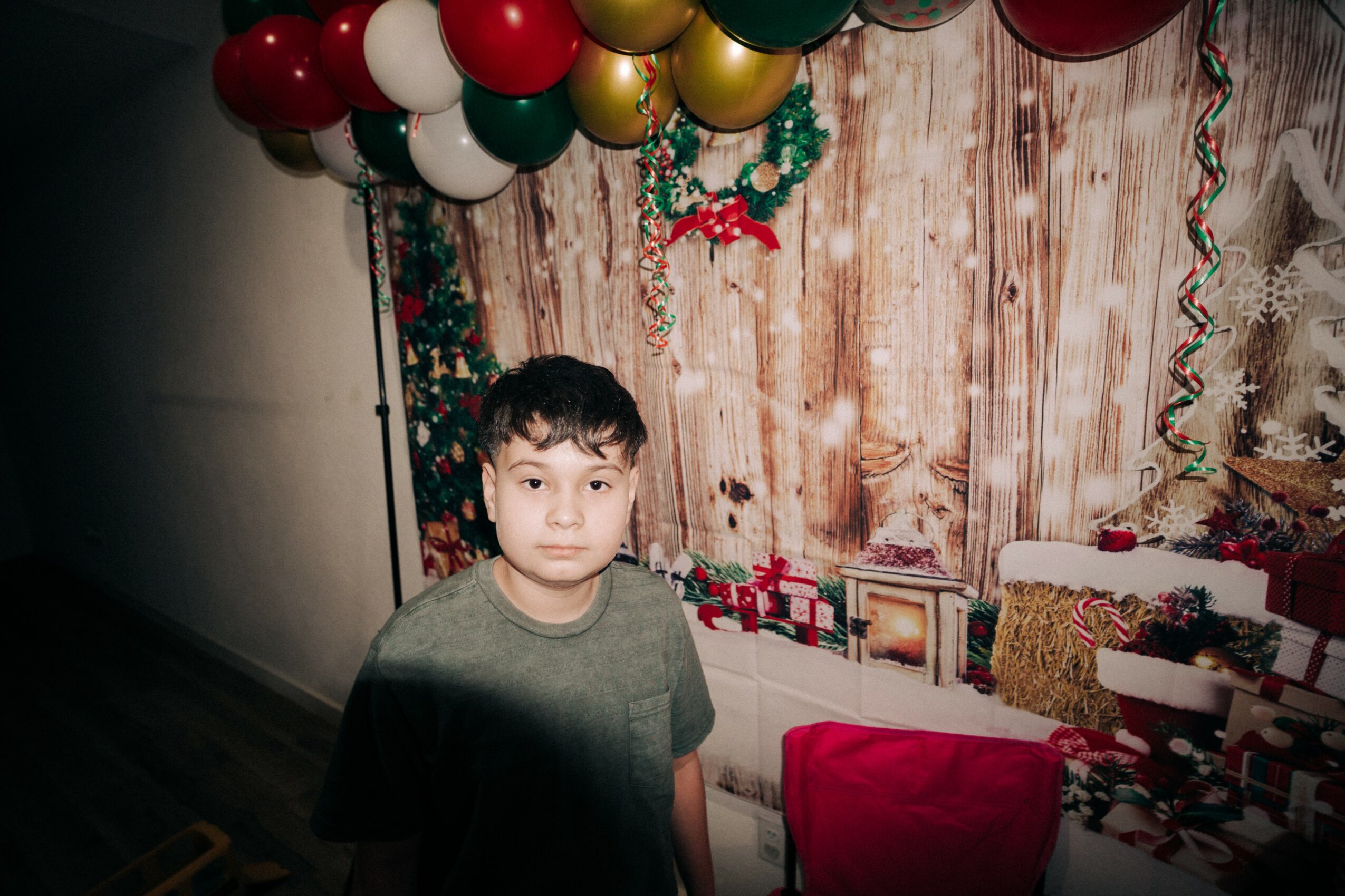 A boy stands in front of a decorated background with balloons and Christmas decorations, looking at the camera.