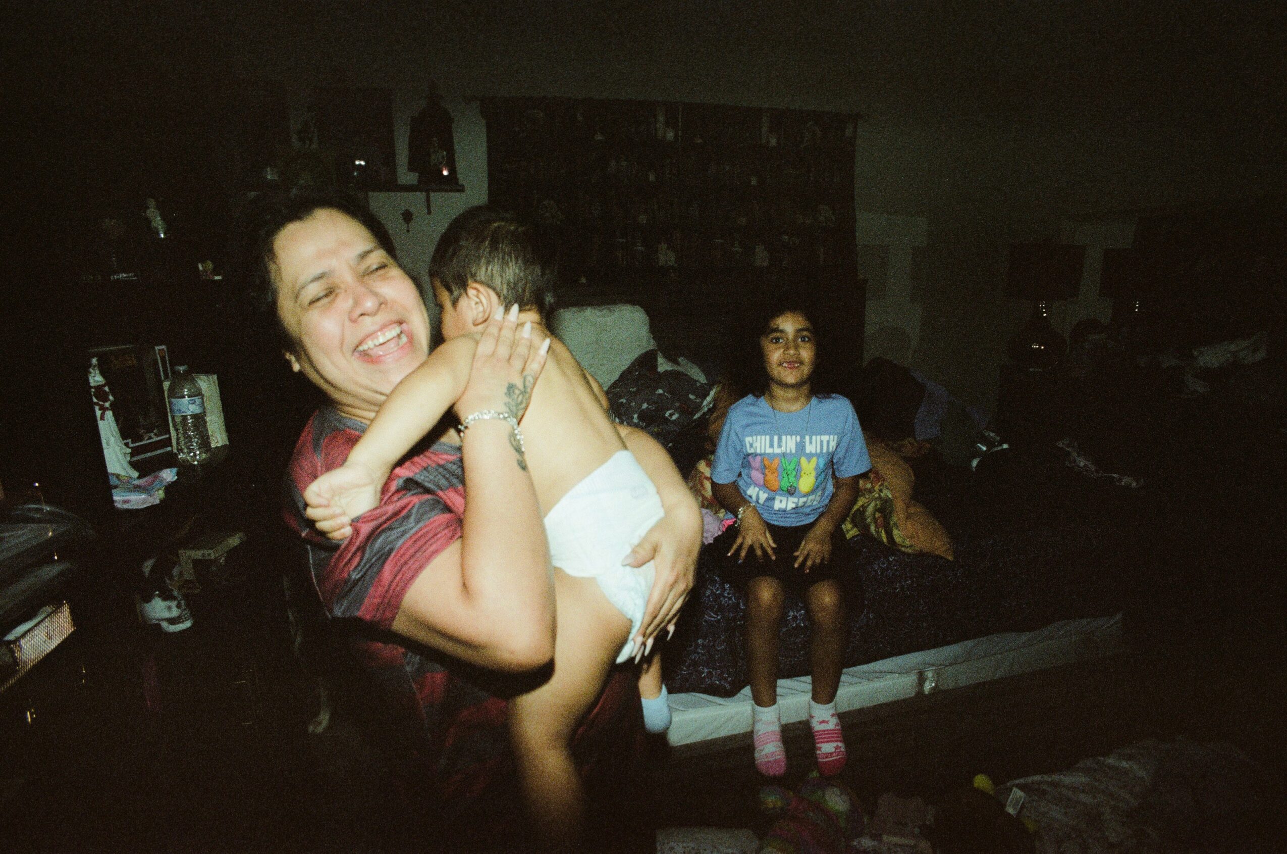 Woman smiling and holding a child, another child sitting on a couch in a dimly lit room.