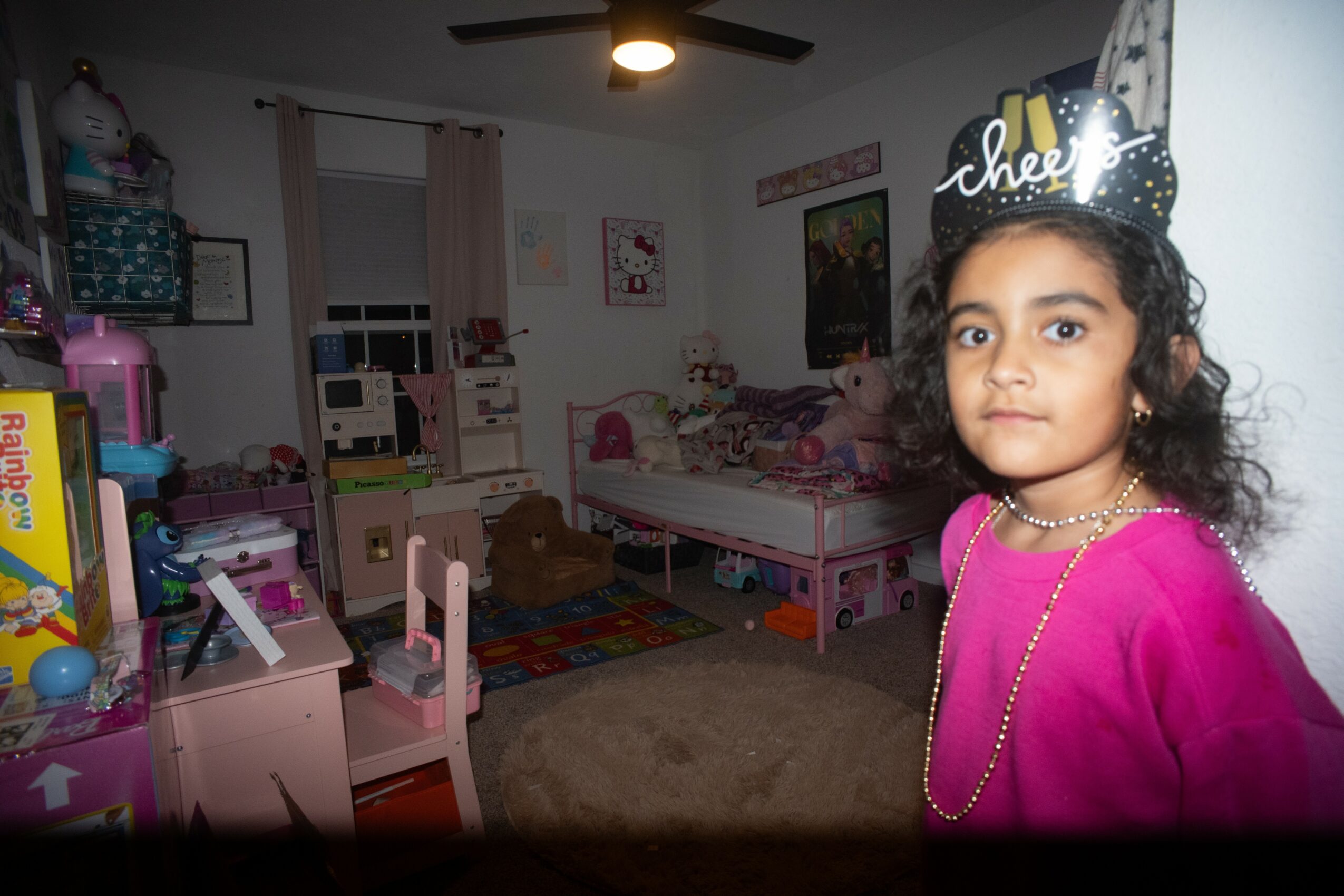 Girl wearing a birthday crown and pink dress stands in a decorated bedroom with pink furniture and toys.