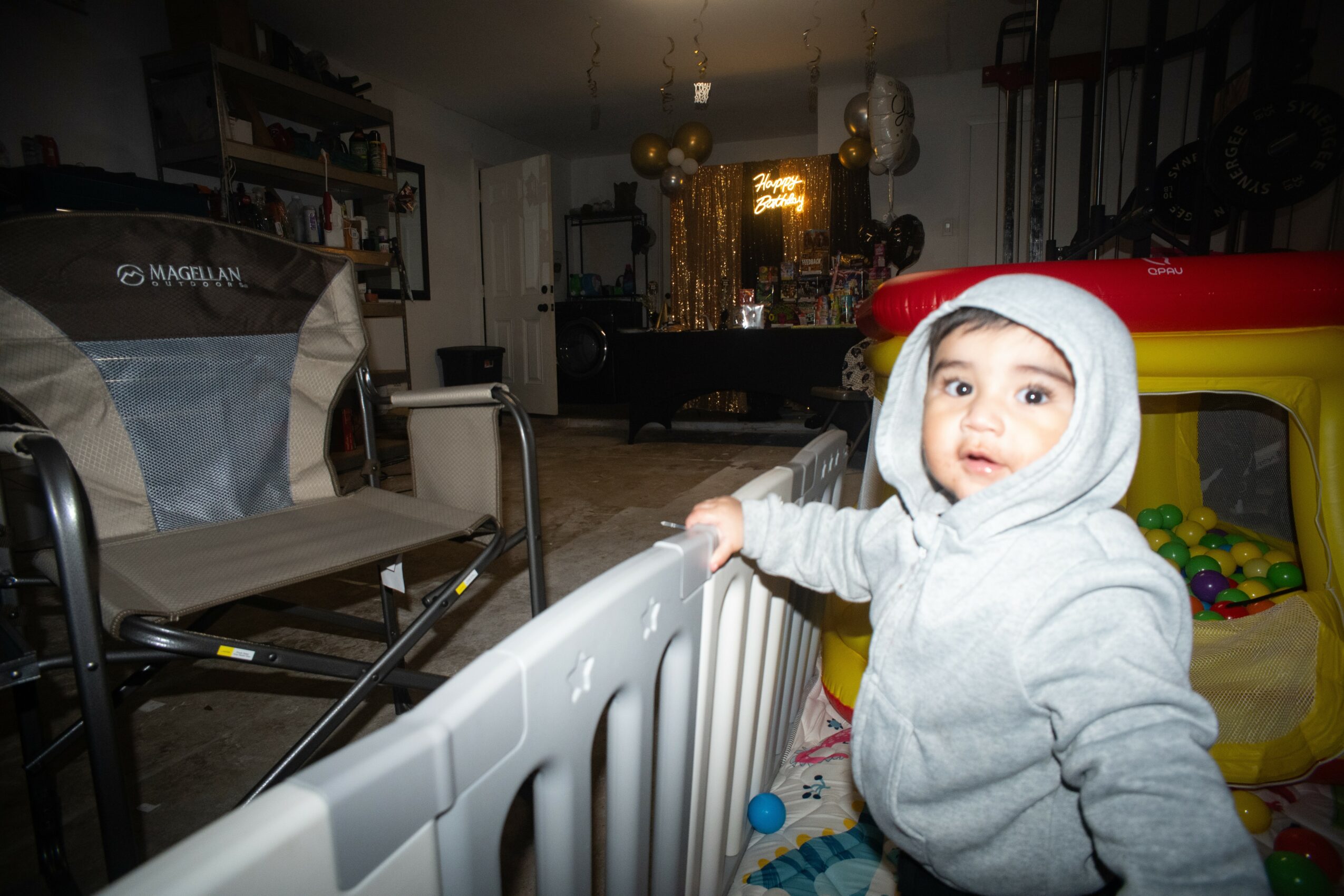 Young child in a gray hoodie standing near a playpen in a cluttered room, with a yellow and red playhouse in the background.