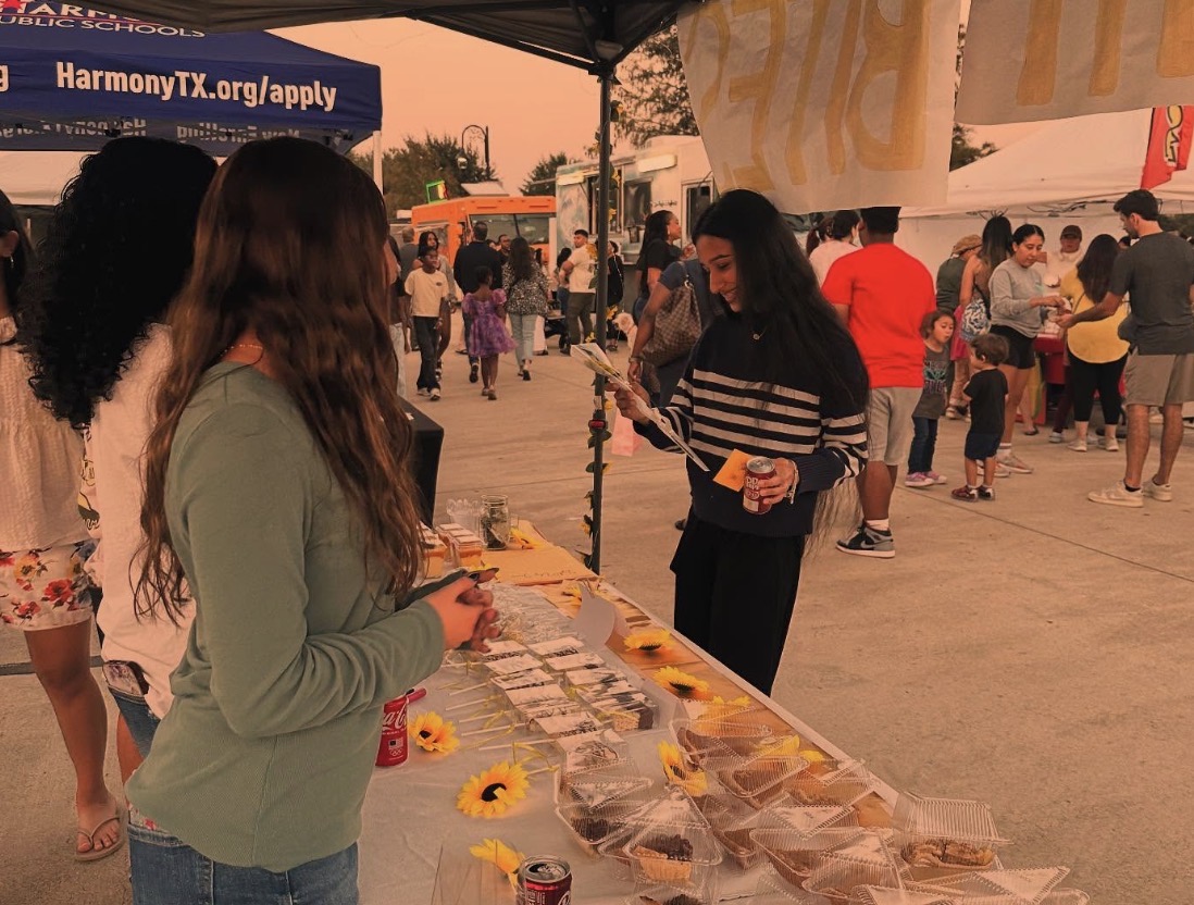 Two women at a table with small items and flowers, outdoors at a busy event with tents and people.