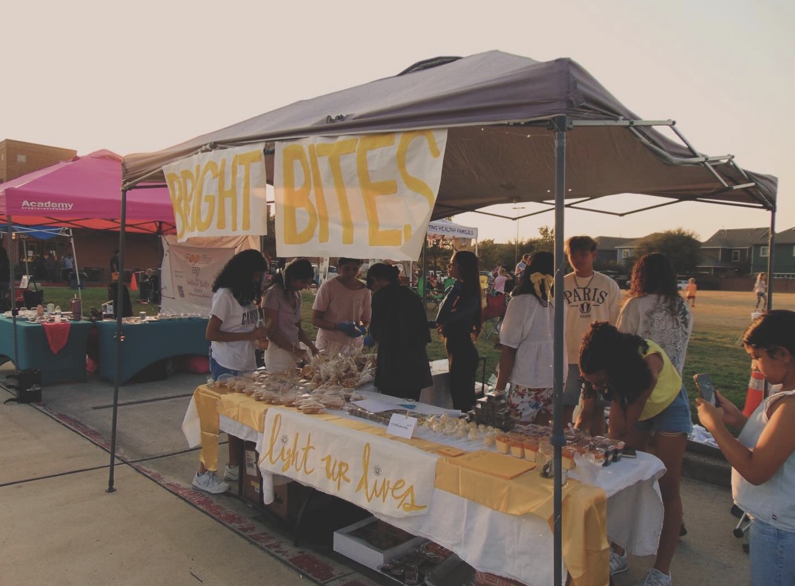 People gather under a tent at a bake sale with signs reading