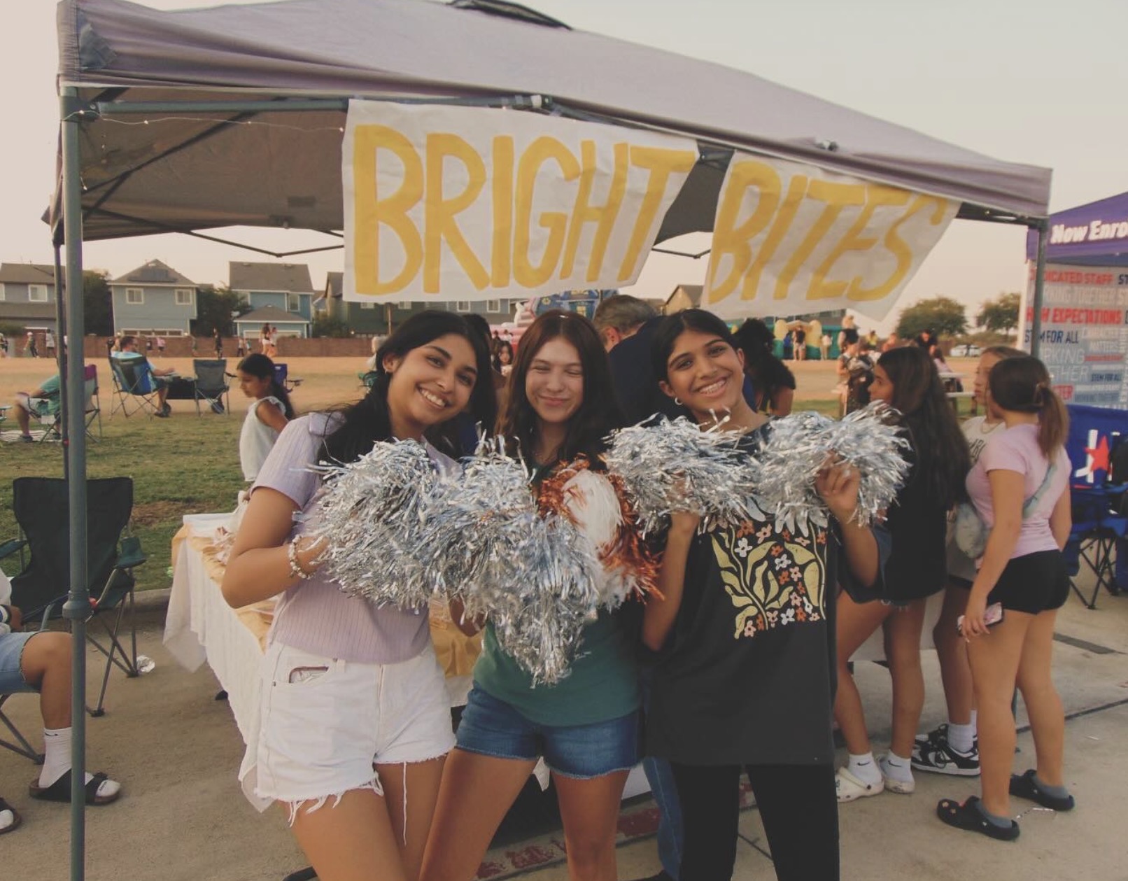 Three smiling women holding silver tinsel, standing in front of a tent with a yellow sign reading 'BRIGHT BITES' at an outdoor event.