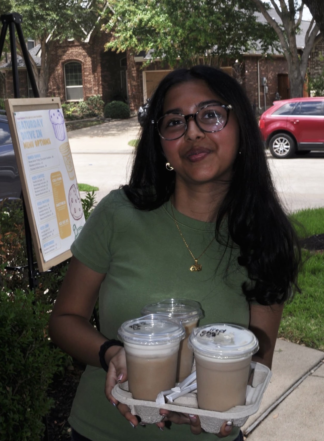 Young woman with glasses and long dark hair holding two iced coffee drinks outdoors, smiling, with trees and a red car in background.