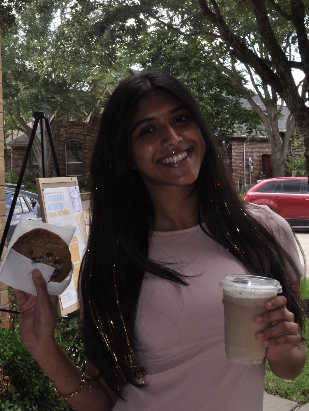 Smiling woman holding a drink and a pastry outdoors with trees and cars in background.