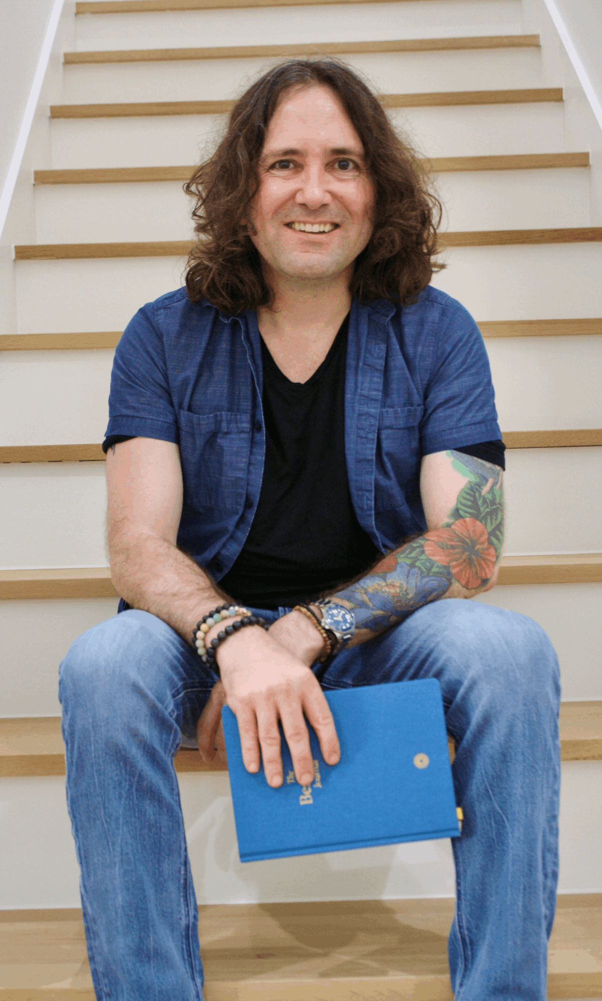 Person with long curly hair sitting on stairs, smiling, wearing a blue shirt and jeans, holding a blue book.
