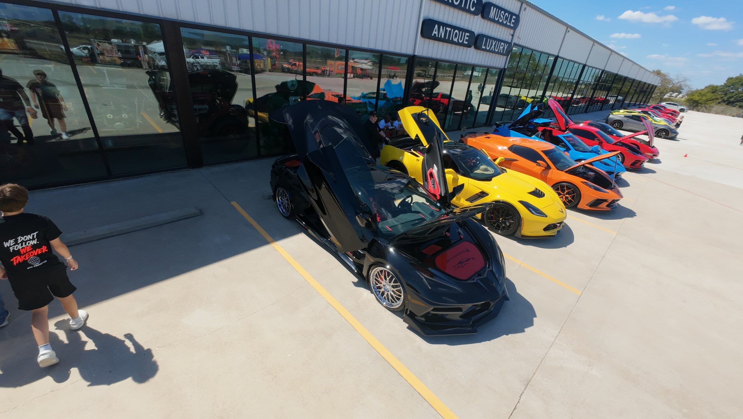 Black sports car parked outside a building with glass windows, several colorful sports cars lined up nearby, and two children walking.