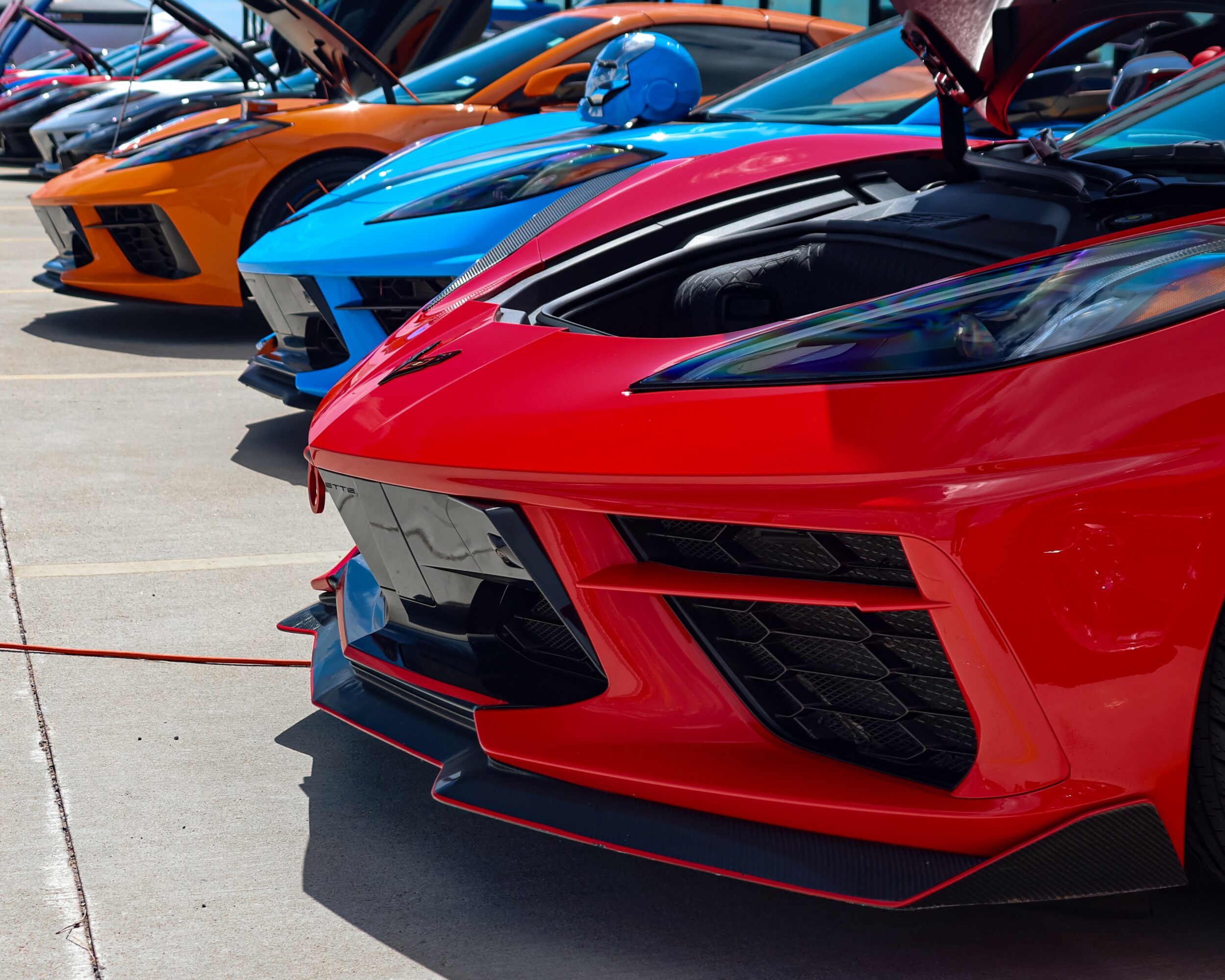 Lineup of colorful sports cars parked outdoors, focusing on front sections and hoods, with a red car in foreground.