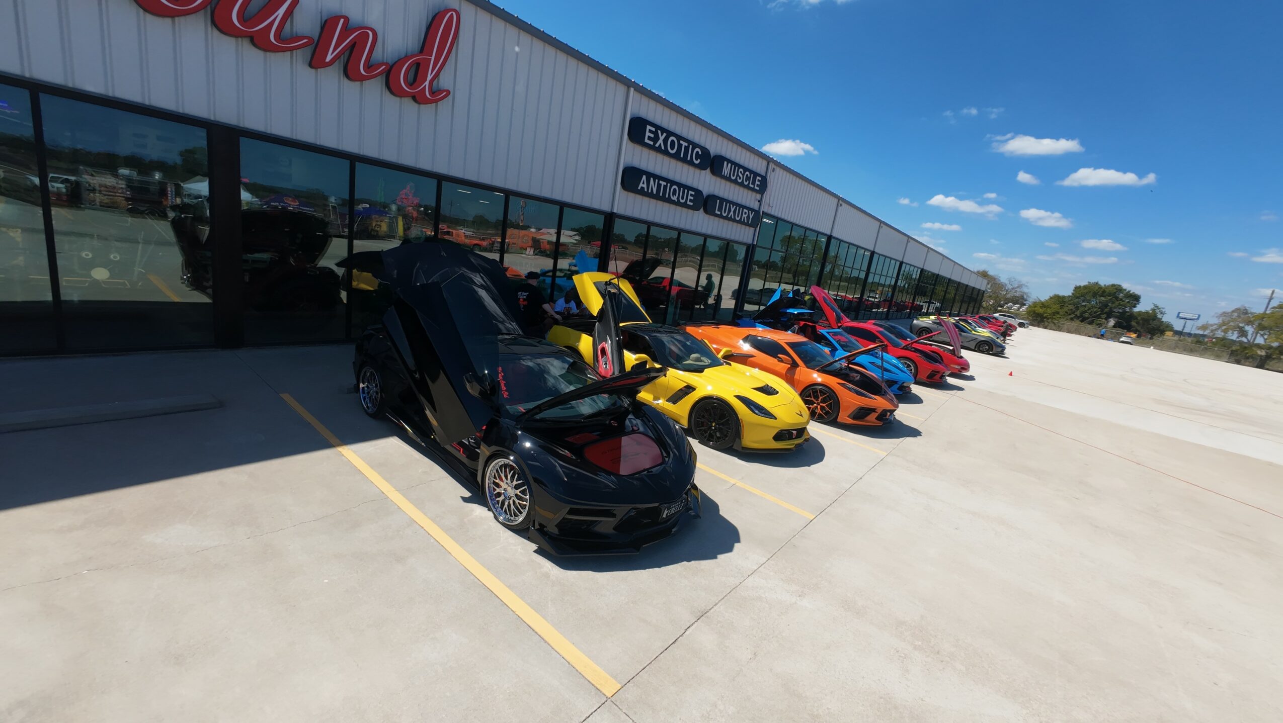 Line of sports cars parked outside a building with large windows and a sign, under a blue sky.
