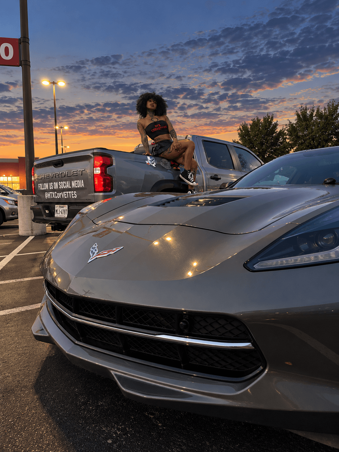 Person sitting on the back of a pickup truck in a parking lot during sunset, with a sports car in foreground.