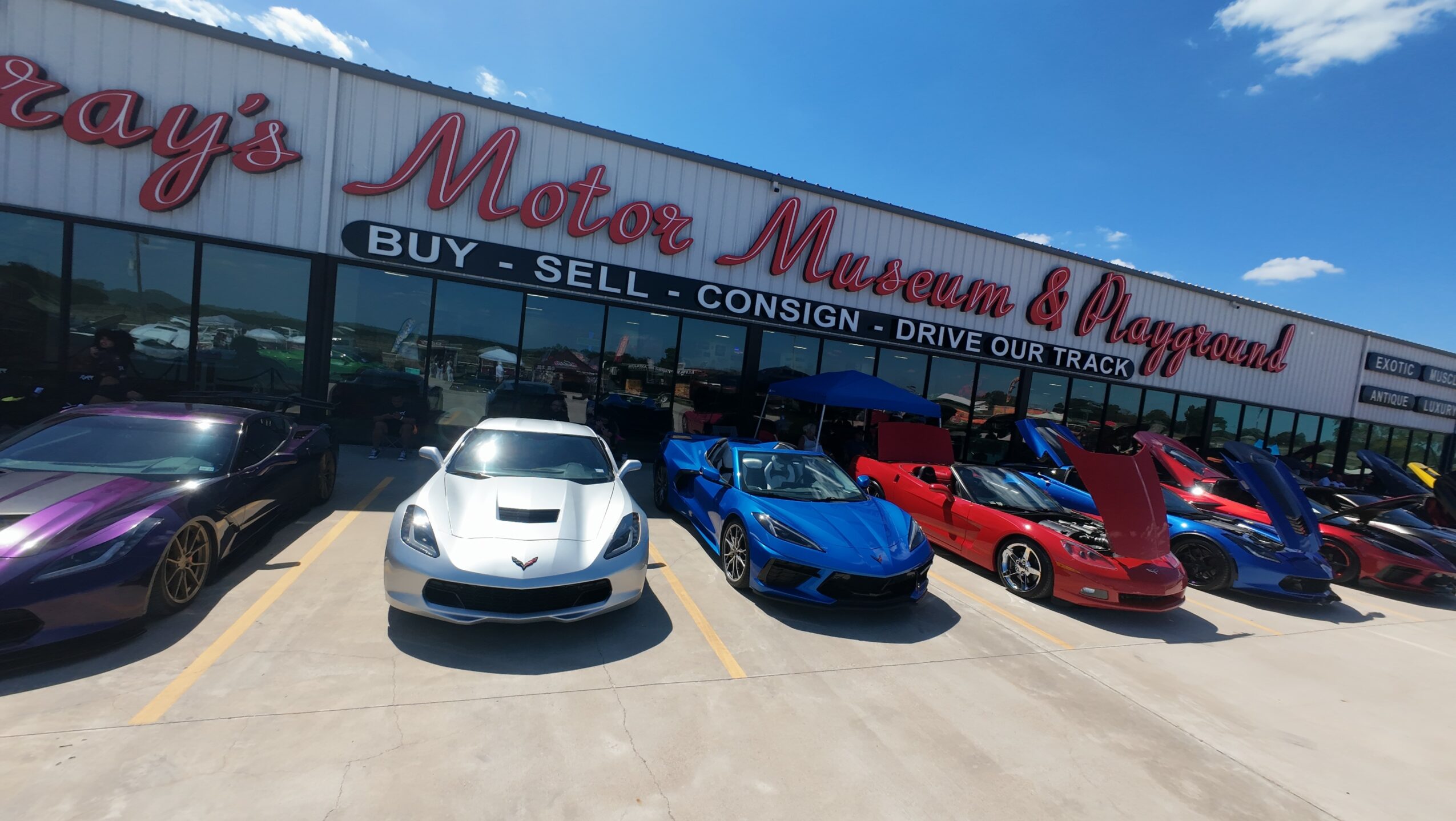 Car dealership with various sports cars parked outside, building signage visible, clear sky overhead.