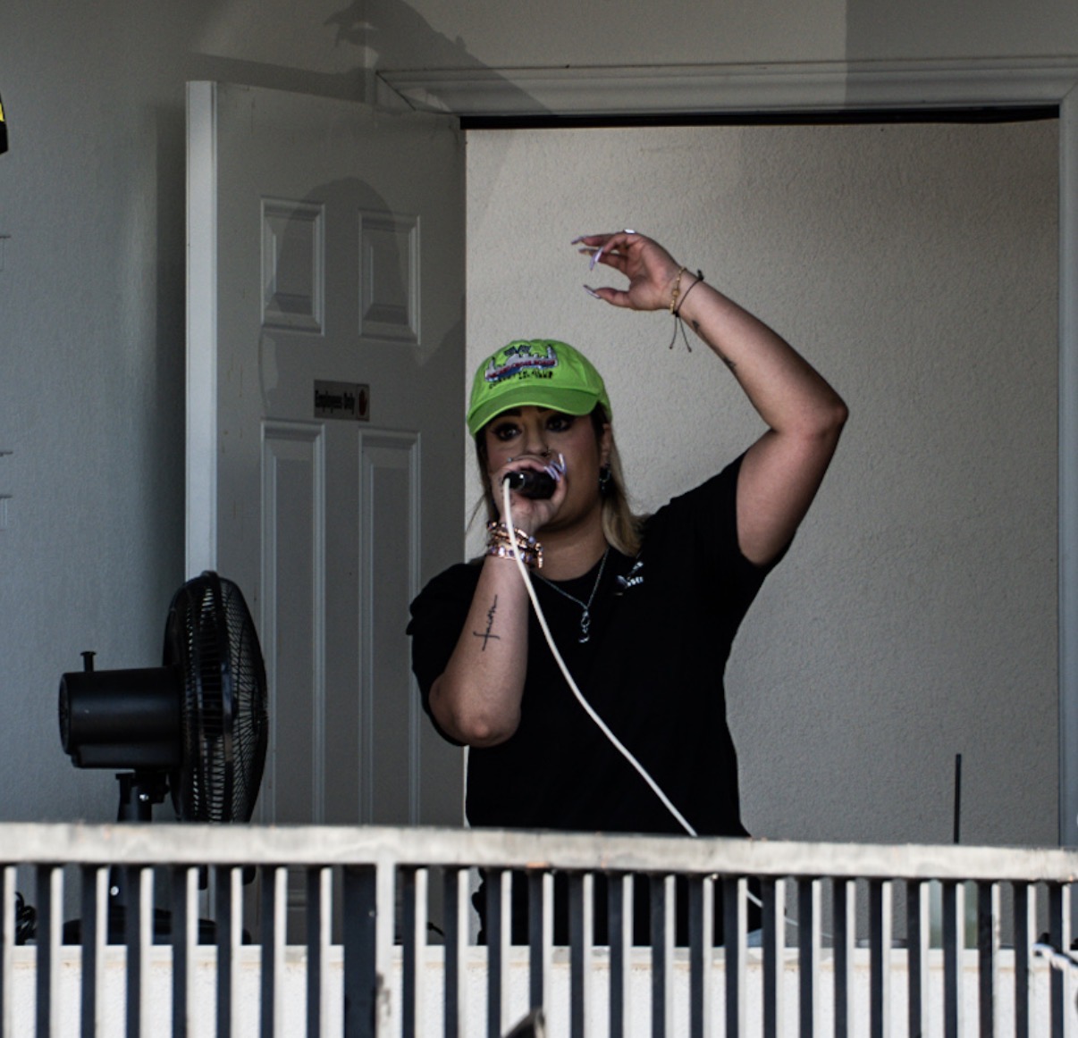 Person wearing a green cap and black shirt, holding a microphone, raising one arm, standing in front of a white wall.