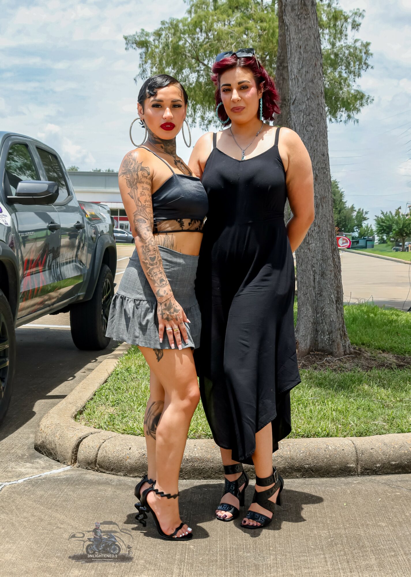 Two women standing outdoors near a tree and parked cars, one in a black crop top and skirt, the other in a black dress.