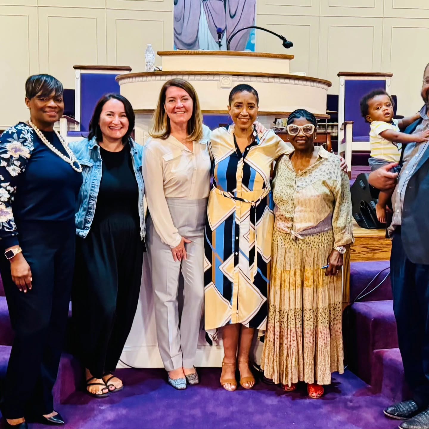 Group of six women standing in a church, smiling, with a pulpit and purple carpet behind them.