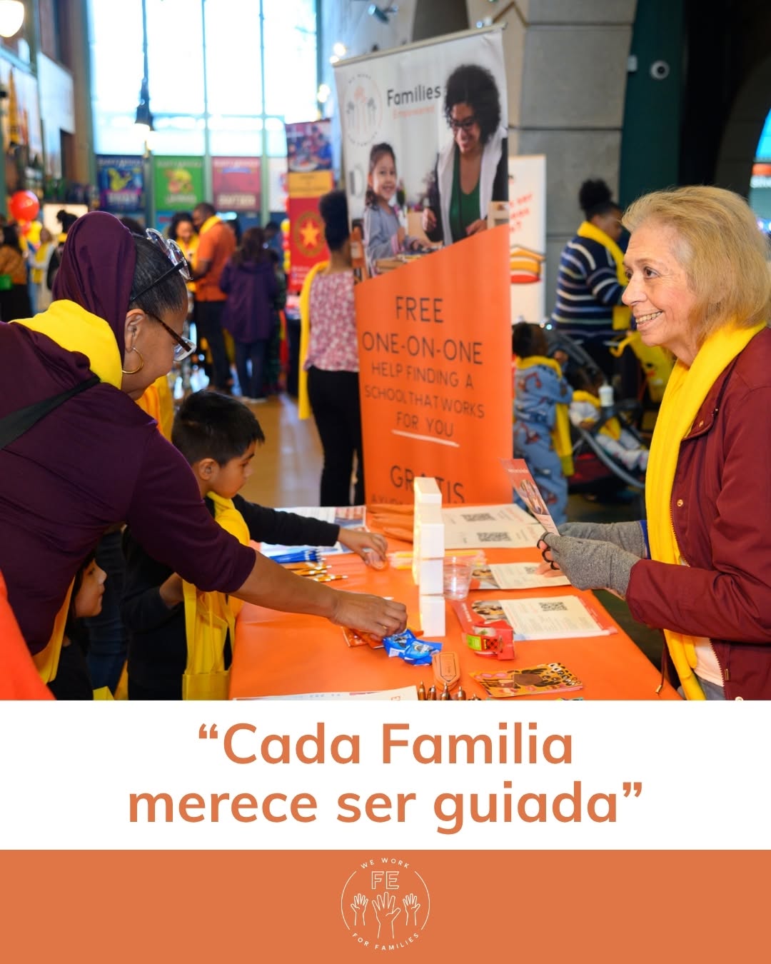 People at a table with brochures and a woman smiling, in an indoor event space with banners and children present.