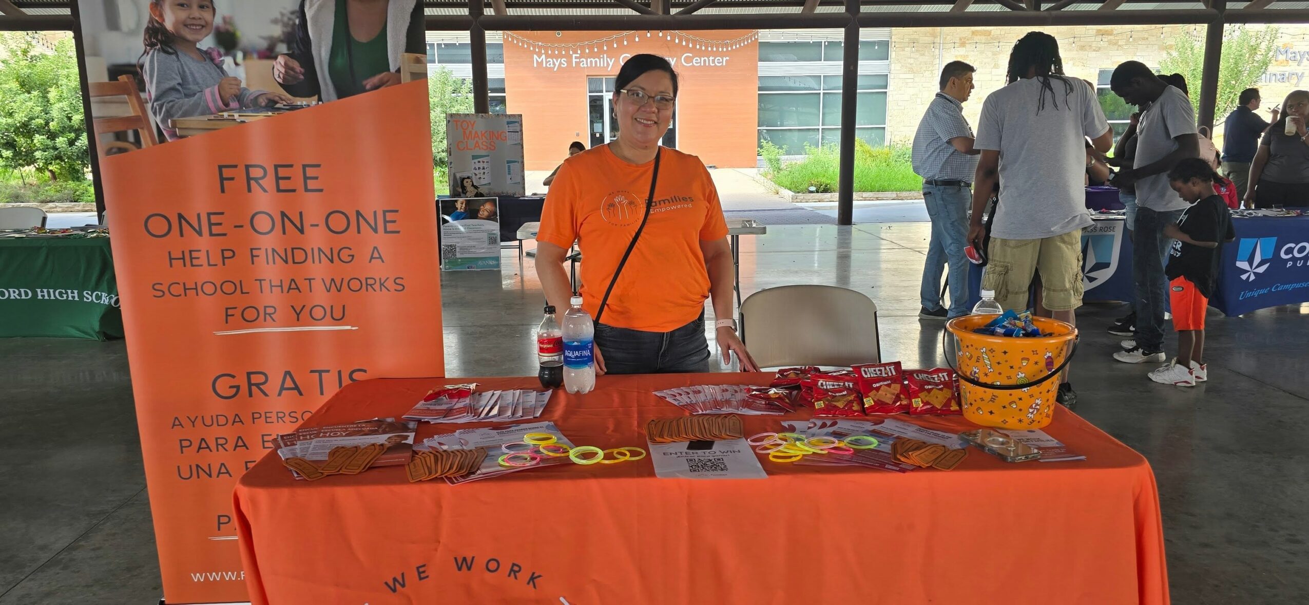 Person standing behind a table with red tablecloth at an outdoor event, with a large orange sign and people in background.