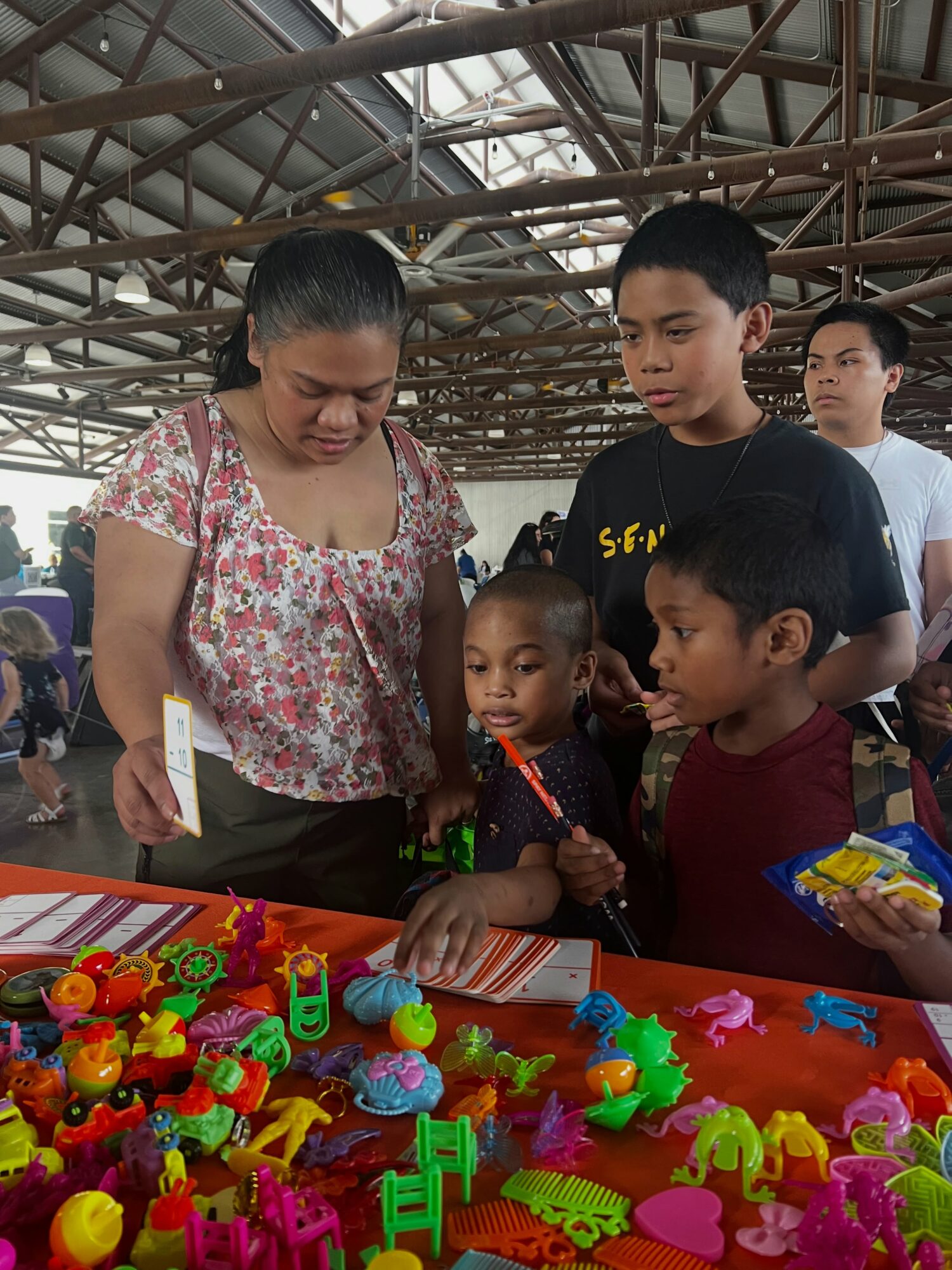 Group of children and a woman looking at colorful toys on a table under a metal roof structure.