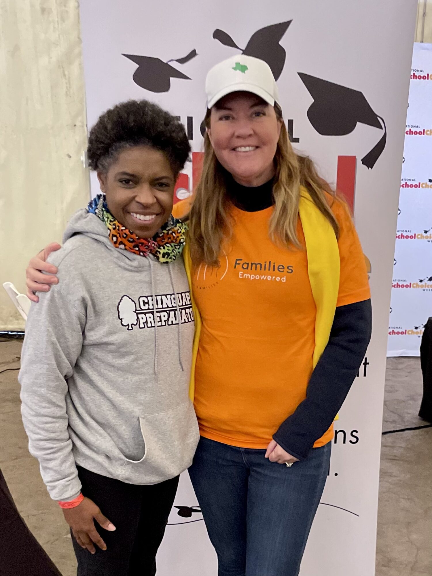 Two women smiling, one wearing a gray hoodie and the other in an orange shirt and white cap, standing in front of a banner.