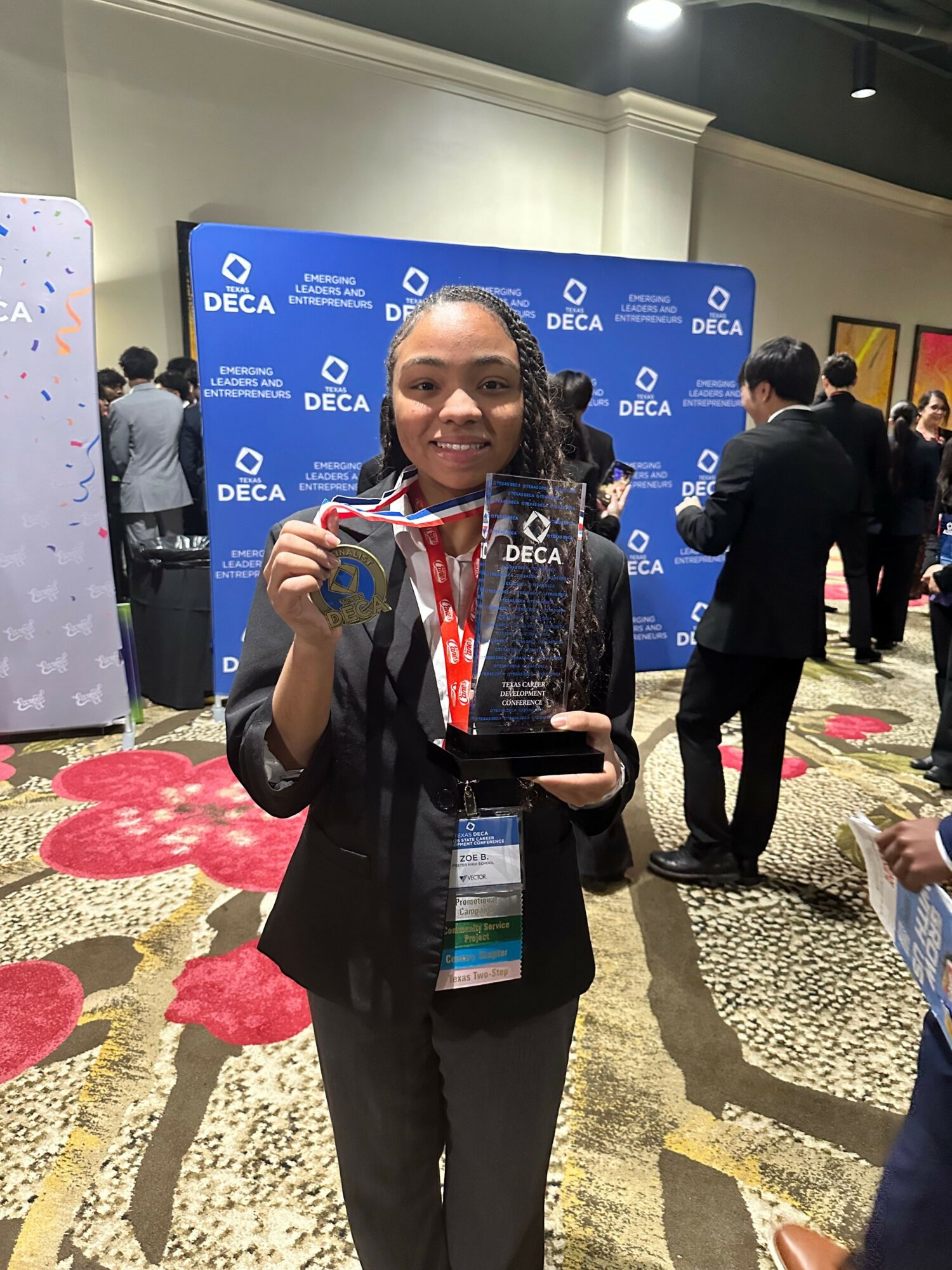 Woman holding a phone and smiling in front of a blue conference backdrop with logos, wearing a black blazer and red lanyard.