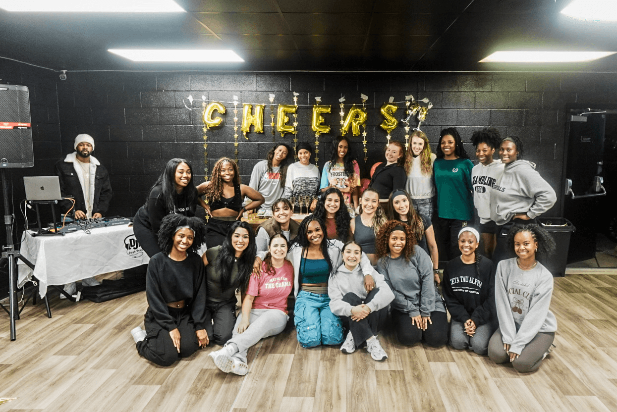 Group of young women and girls posing in front of a black wall with gold 'CHEERS' balloons, smiling and standing together.